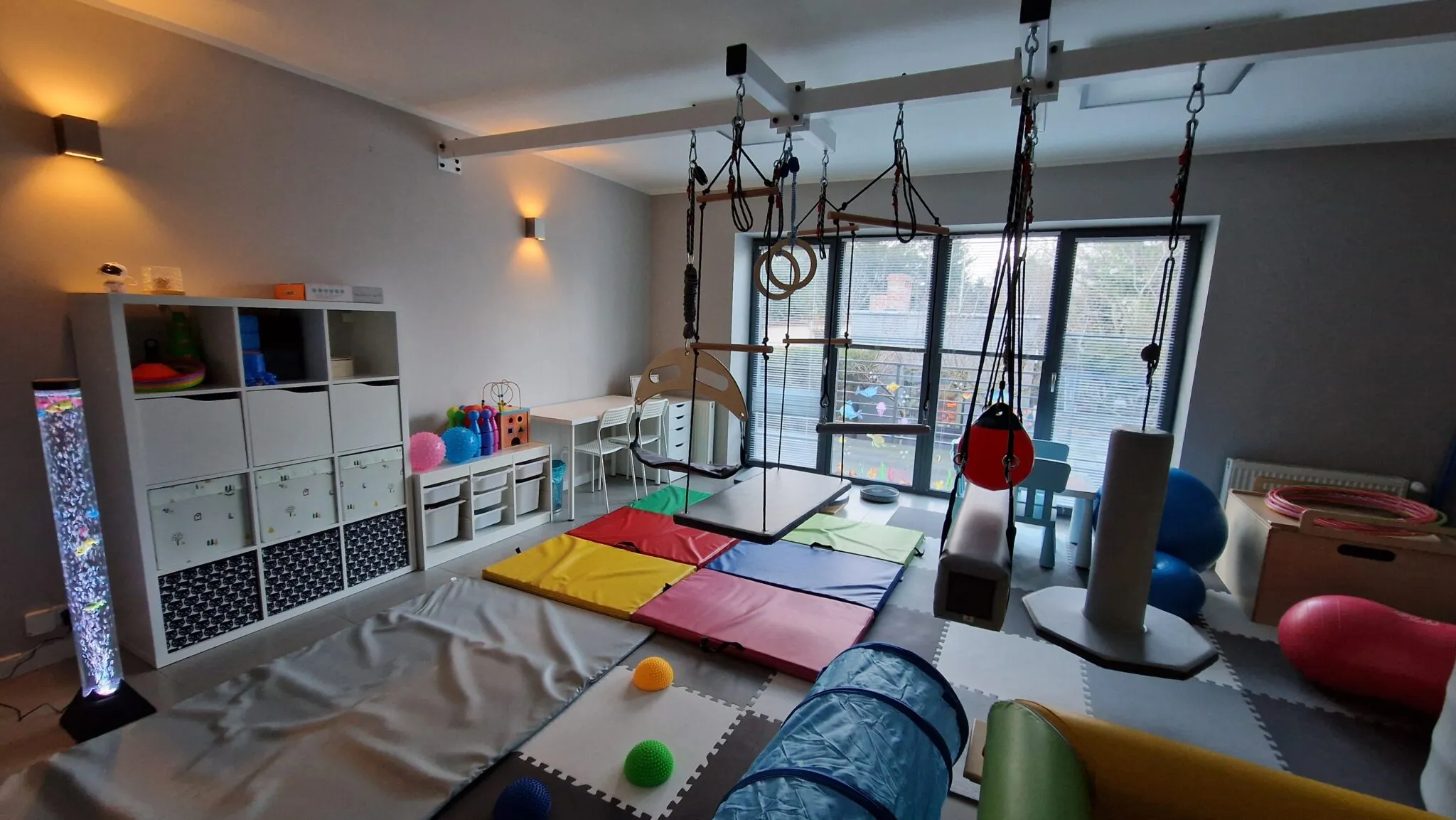 Sala integracji sensorycznej z regałem na pomoce, kolumną wodną, kolorowymi materacami, tunelem i huśtawkami